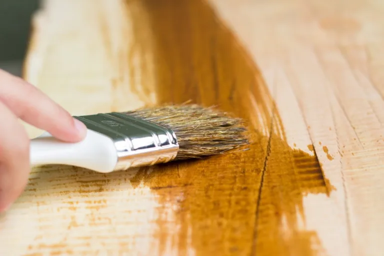 A imagem mostra uma mão aplicando stain com um pincel sobre uma superfície de madeira, destacando a textura e o brilho do acabamento.